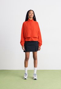 Rød zip-up jakke med elastiske mansjetter, kombinert med en svart skjørt. Modellen har på seg hvite sokker og grå joggesko, stående på et lys grønt gulv.