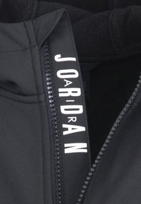 Veste noire zippée avec doublure en polaire ; présente un logo blanc bien en vue sur la bande de fermeture éclair ; texture du tissu lisse.