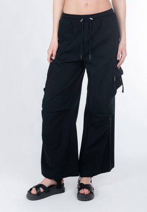 Zwarte cargobroek met een ontspannen pasvorm, trekkoord in de taille, zijzakken en een gladde textuur. Gecombineerd met zwarte sandalen met bandjes.