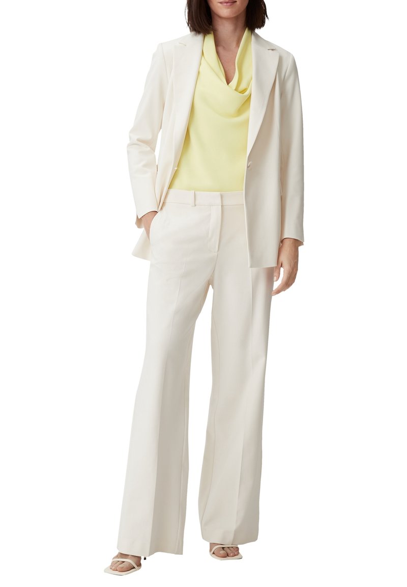 Femme portant un costume crème avec un pantalon à jambes larges et un blazer sur un top sans manches jaune clair, ainsi que des sandales ouvertes crème.