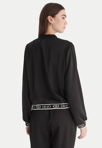 Giacca bomber nera in satin con maniche lunghe, polsini e vita elasticizzati, caratterizzata da una banda con logo bianco. Tessuto liscio e silhouette classica.