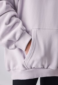 Lyse lilla hættetrøje med ribbede manchetter og kant, der har en frontlomme og blødt, tekstureret stof. Close-up billede af ærme og lomme.