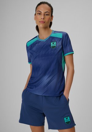 Kobieta ubrana w niebieski i turkusowy strój sportowy z logo 4F na koszulce i szortach, stojąca z rękami w kieszeniach na jednolitym tle.
