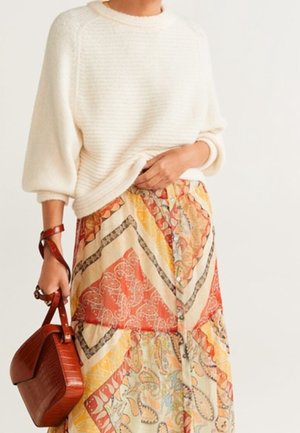 Pull en maille couleur crème avec un tissu texturé, associé à une jupe maxi à motifs avec des dessins géométriques orange et jaune. Tient un sac à main marron.