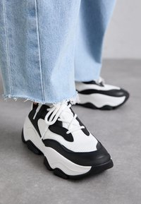 Sorte og hvite joggesko med teksturert mesh og glatt gummimateriale. Kraftig såle, fremtredende snøring og et moderne design.