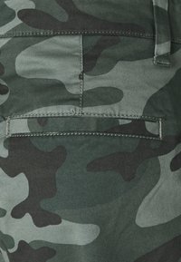 Kamouflageväv i nyanser av grönt, svart och grått. Strukturerad yta med fickor och kontrasterande sömdetaljer.