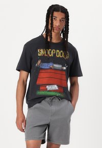 T-shirt noir en coton avec un graphique coloré d'un chien de dessin animé sur une niche rouge. Porté avec un short gris, mettant en valeur une coupe décontractée.