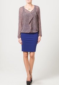 Blouse transparente à motifs avec manches longues, associée à une jupe bleue ajustée mi-longue. Des chaussures noires avec un accent coloré complètent la tenue.