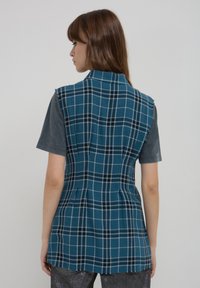 Gilet a scacchi turchesi con un design strutturato, caratterizzato da un colletto e una linea senza maniche. Tessuto testurizzato con quadri neri a contrasto e accenti grigi.