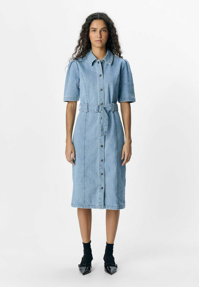 Vestido de camisa de mezclilla azul claro con mangas cortas, cierre de botones y cintura ajustada con cinturón; presenta un corte recto y un dobladillo a la altura de la rodilla.