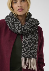 Black and gray patterned scarf with fringed ends, made from soft material, worn over a dark green top beneath a burgundy coat.