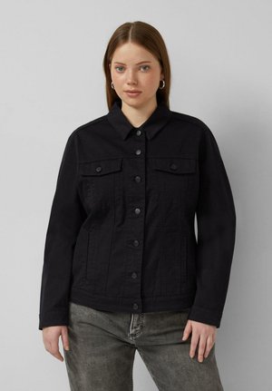 Junge Frau mit langen braunen Haaren, die silberne Creolen, eine schwarze Jacke mit Knöpfen und ausgewaschene graue Jeans trägt, steht vor einem grauen Hintergrund.