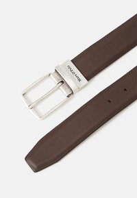 Ceinture en cuir marron avec une boucle en métal argenté. La boucle a une forme rectangulaire avec un logo gravé. Comprend cinq trous de réglage.