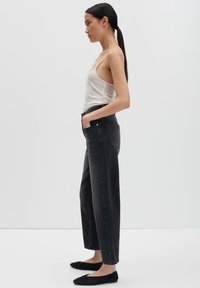 Vrouw in zijaanzicht, gekleed in een witte spaghettibandtop, hoge zwarte jeans en zwarte platte schoenen, tegen een effen achtergrond.