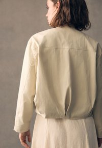 Lichtbeige jas met een gladde textuur, voorzien van een relaxte pasvorm, verlaagde schouders en een naad langs de achterkant. Draagt over een textiel lichtgekleurde kleding.