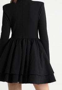 Musta mekko, jossa on tiukka vartalonmyötäinen yläosa ja pitkät hihat, ja joka sisältää kerroksellisen, levenevän hameen, jossa on röyhelöyksityiskohtia ja sileä pinta.