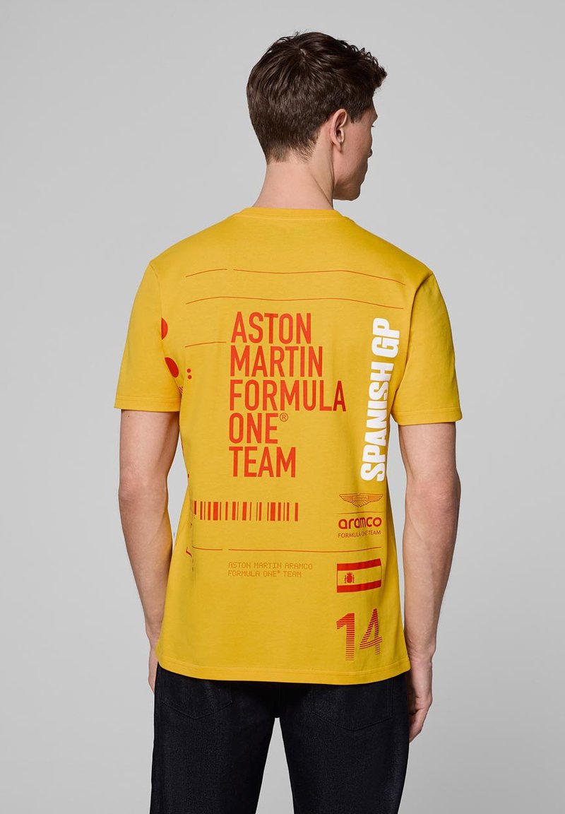 Żółta koszulka z czerwonymi napisami i grafikami: "Zespół Formuły 1 Aston Martin", "GP Hiszpanii", z elementami projektu w postaci kodu kreskowego i flag.