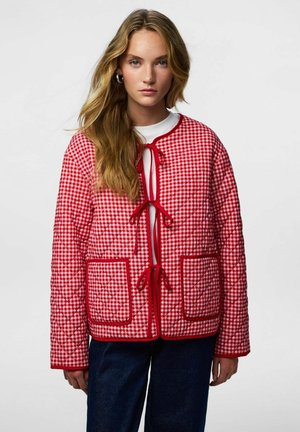 Žena s dlouhými blond vlasy nosí červenobílou prošívanou bundu károvaného vzoru se zavazováním vpředu a velkými náprsními kapsami, doplněnou tmavými džínami.