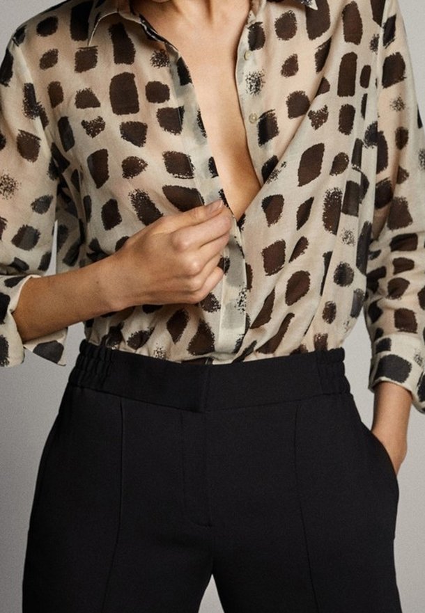 Personne portant un chemisier beige à motifs avec des taches carrées foncées et un pantalon noir taille haute, boutonnant le chemisier d'une main.