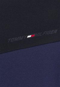 Tissu bleu marine à texture lisse avec le logo "TOMMY HILFIGER" en gris foncé au-dessus d'une bande horizontale rouge, blanche et bleue.