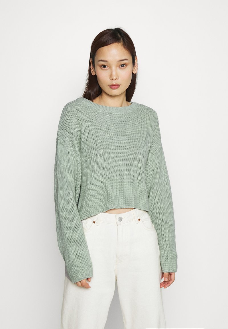 Femme aux cheveux raides et foncés portant un pull en tricot vert clair et un pantalon blanc taille haute sur fond blanc uni.
