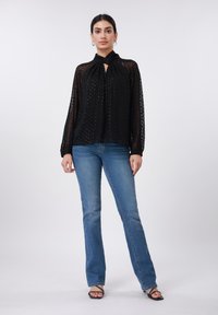 Blouse noire transparente avec texture à pois, col montant avec détail à nouer, manches longues. Associée à un jean évasé bleu clair et des sandales noires.