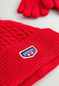 Röd stickad mössa med en "Superdry" logotypapplikation och matchande röda handskar på en vit yta.