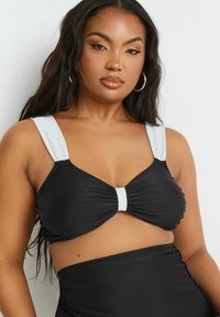 Svart baddräktstopp med breda vita axelband och en tvistad frontdetalj. Den släta texturen framhäver den figurnära designen, med matchande högmidjade bottoms.