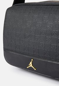 Svart teksturert veske med preget "23"-mønster, glatte lærdetaljer og en gull Jumpman-logo. Glidelås lukking på toppen.