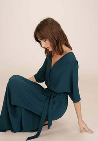 Teal wikkeljurk met korte mouwen, diepe V-hals en een strik in de taille. Vloeiende stof creëert een zachte silhouet; vloerlang met een subtiele textuur.