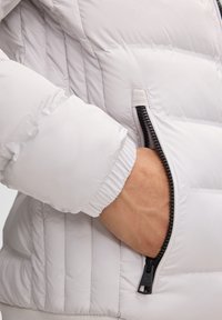 Hellgraue Puffy-Jacke mit gestepptem Design, elastischen Bündchen und einem schwarzen Reißverschlussdetail an einer Seitentasche.