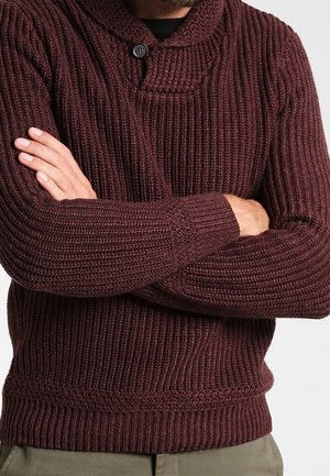Homme portant un pull en maille bordeaux foncé avec un col châle et un bouton, les bras croisés sur la poitrine.