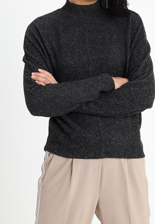 Personne portant un pull gris foncé à manches longues, les bras croisés, et un pantalon beige avec des bandes blanches sur les côtés.
