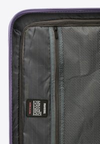 Interno della valigia viola con tasche in rete grigia, scomparti con zip e etichetta di cura. Tessuto texturizzato con elementi a strisce.