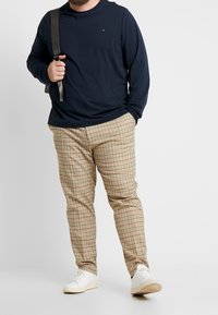 Granatowa koszula z długim rękawem z małym logo, połączona z beżowymi spodniami w kratę z czerwonymi i czarnymi akcentami; białe trampki; szara torba na ramię.