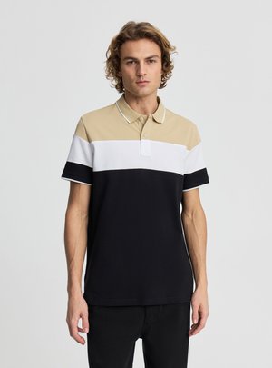 Giovane uomo che indossa una polo a blocchi di colore beige, bianco e nero, in piedi davanti a uno sfondo chiaro e semplice.