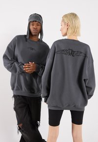 Felpa grigia oversize con una texture liscia, caratterizzata da una piccola grafica di un'ala sul petto e un design di un'ala più grande sulla schiena.