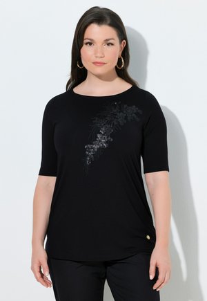 Frau trägt ein schwarzes Kurzarmtop mit paillettiertem Blumenmuster auf der Brust und goldenen Creolen, steht vor einem weißen Hintergrund.