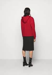 Red hooded sweatshirt made of cotton, paired with a black knee-length skirt featuring a slit, and black heeled ankle boots.