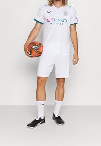 Bílý fotbalový dres Puma s tyrkysovými akcenty, v kombinaci s bílými šortkami a černými kopačkami. Drží oranžovým a černým fotbalovým míčem.