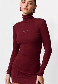 Bordowa prążkowana sukienka z golfem, długimi rękawami i dopasowaną sylwetką. Na piersi znajduje się biały napis "LAMANIA". Gładka faktura.