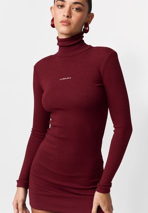 Bordowa prążkowana sukienka z golfem, długimi rękawami i dopasowaną sylwetką. Na piersi znajduje się biały napis "LAMANIA". Gładka faktura.