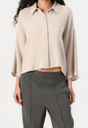 Kvinde iført en cremefarvet, tekstureret bluse med knapper og brede ærmer samt mørkegrå, stribede bukser, stående mod en ensfarvet baggrund.