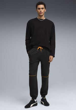 Joven de pie, vistiendo una camiseta negra de manga larga, pantalones jogger negros con cordón y ribete naranja, y zapatillas Puma negras.