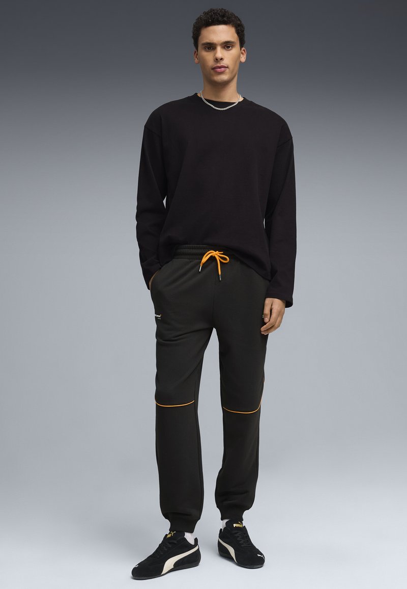 Joven de pie, vistiendo una camiseta negra de manga larga, pantalones jogger negros con cordón y ribete naranja, y zapatillas Puma negras.