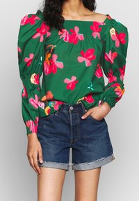 Blouse verte à fleurs avec manches bouffantes, ornée de motifs de fleurs roses et jaunes. Associée à un short en denim foncé, retroussé à l'ourlet.