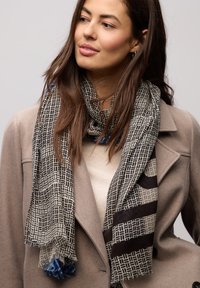 Sort-hvidt mønstret halstørklæde med frynser, prydet med geometriske designs. Båret over en beige top og en grå frakke.