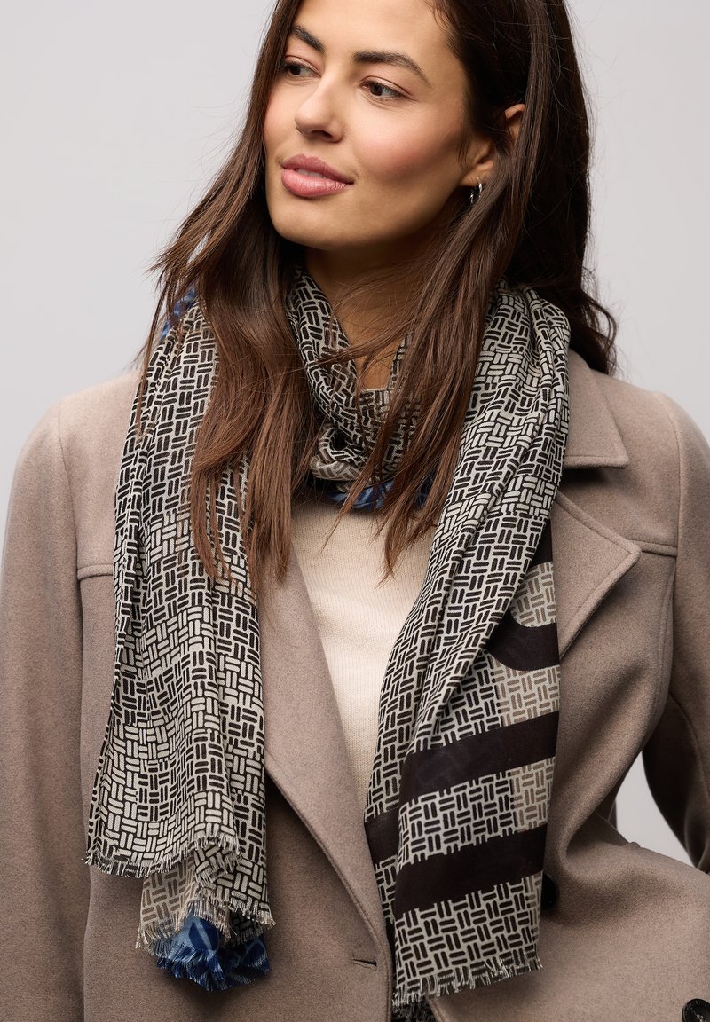 Sort-hvidt mønstret halstørklæde med frynser, prydet med geometriske designs. Båret over en beige top og en grå frakke.