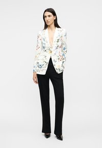 Nő, aki fehér, virágos blézert visel színes mintákkal, fekete bőszárú nadrágot és fekete hegyes orrú cipőt, egyik kezét zsebében tartva áll.