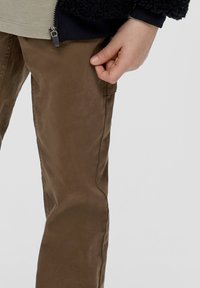 Bruine katoenen broek met een gladde textuur, voorzien van een zijzak en een getailleerd silhouet. Hand zichtbaar bij de taille, met een neutraal shirt erboven.